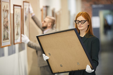 Red haired art manager in eyeglasses looking at painting in her hands while standing at art gallery...