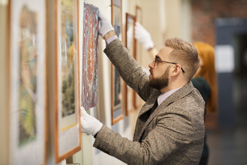 Handsome bearded man in eyeglasses and in suit hanging the picture on the wall and preparing for...