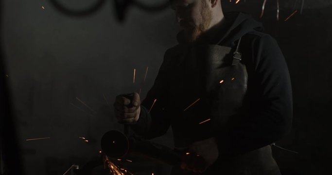Portrait of a blacksmith perfecting his blade in a dark interior of his metal workshop. 