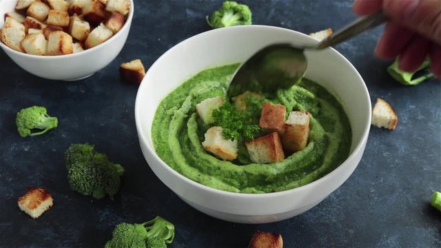 hand with tablespoon mixing broccoli puree soup with croutons first person view