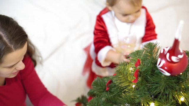 baby and pregnan mom hang red ball toy on Christmas tree. happy childhood concept. child and mother decorate tree with christmas balls. small child and a parent are playing by Christmas tree.