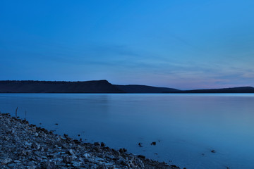 long exposure landscape of ocean bay peaceful smooth water surface stone shore line foreground and horizon main land silhouette background in calm summer time weather twilight evening lighting