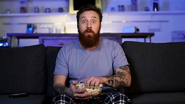 Excited bearded man sitting on the couch, eating popcorn and watching tv (or football). Slowmotion.
