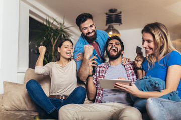 Four casual happy friends laughing shopping online together in a tablet sitting on a sofa in the living room at home