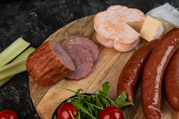 on a dark table a wooden breakfast board with a suggestion of snacks: cheese, salad, tomatoes, sausages