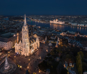 Naklejka premium Budapest, Hungary - Aerial drone view of the beautiful illuminated Matthias Church at blue hour with Fisherman's Bastion (Halaszbastya), Margaret Bridge and Parliament of Hungary at background
