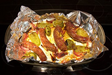 Roasted sausages with onions and peppers in a glass baking pan into aluminum foil on a black ceramic cooktop