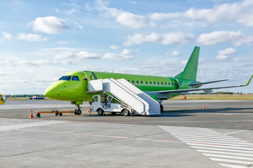 Modern green civil passenger aircraft parked on airfield. A ladder is docked to the plane. Airplane waiting for passengers. Blue sky. Vacation and traveling concept. Copy space. Air transportation.