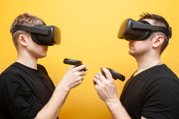 two guys in VR glasses on a yellow background play a shooter, friends gamers rivals in virtual reality glasses fight and shoot