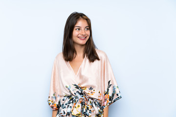 Caucasian girl with kimono over isolated blue background laughing and looking up