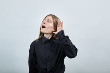 Caucasian young woman in fashion black shirt keeping hand near ear, listening, scream isolated on gray background in studio. People sincere emotions, lifestyle concept.