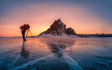 Fotograf trägt rote Kleidung und fotografiert den Schamanka-Felsen bei Sonnenaufgang mit natürlichem brechendem Eis in gefrorenem Wasser am Baikalsee, Sibirien, Russland.