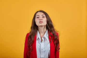 Attractive caucasian young woman in fashion white shirt and red jacket blowing, send air kiss isolated on orange background in studio. People sincere emotions, lifestyle concept.