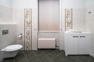 Modern interior of bathroom in light tones with black tile on the floor. Toilet. Dresser. Blinds on the window.