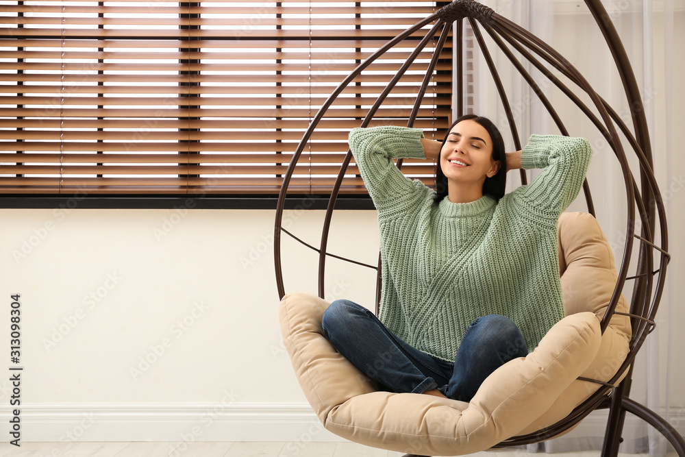 Poster Young woman relaxing in hanging chair near window at home. Space for text