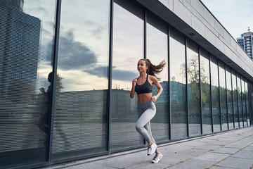 Young athletic woman running in the city, shot of girl runner on the glass building background