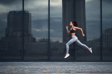 Young athletic woman running in the city, shot of girl jumps on the glass building background
