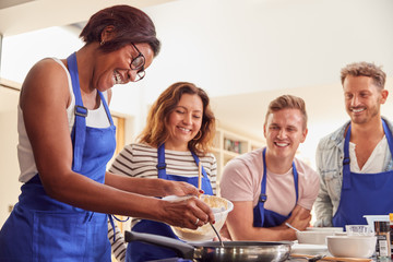 Female Teacher Making Pancake On Cooker In Cookery Class As Adult Students Look On