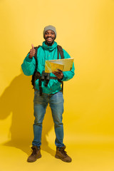 Full length portrait of a cheerful young african-american tourist guy with bag and map isolated on yellow studio background. Preparing for traveling, looking for trip way, smiling and greeting.