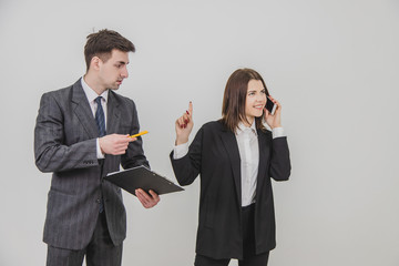 Handsome busy businesswoman is talking on the phone, pointing his finger up for her secretary to wait. Male secretary standing with clipboard, waiting for boss's signature.