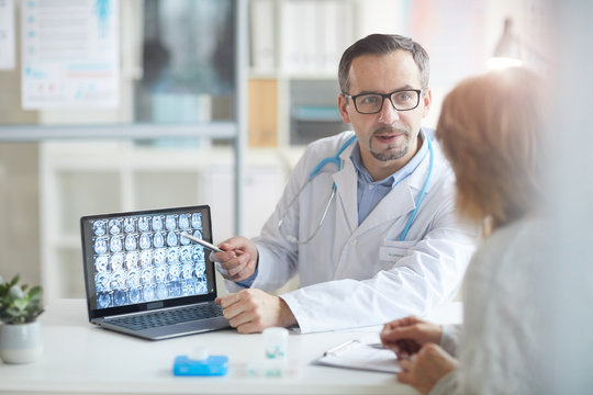 Male doctor explaining the illness to his patient and pointing at laptop with x-ray images while they sitting at the table at office