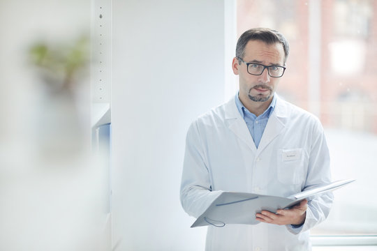 Portrait of mature male doctor in white coat standing and looking at camera while filling the medical card at office