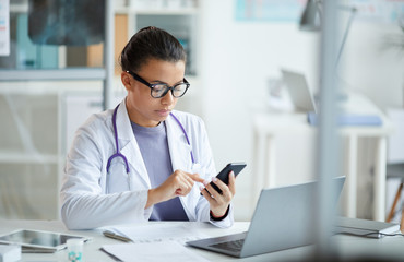 Serious female doctor in white coat sitting at her workplace and typing a message on her mobile...
