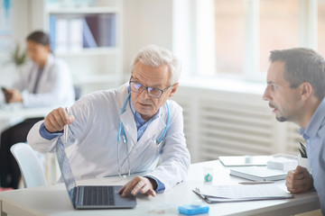 Senior doctor in eyeglasses sitting at the table pointing at laptop computer and discussing illness together with his patient