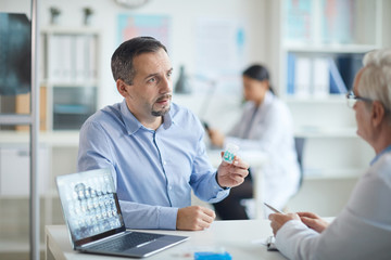 Mature man holding bottle of pills and discussing treatment together with his doctor at the table at office