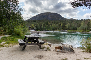 Camping rest area for picnic with wooden table bench on sand by the cold sparkling river in pine forest mountains of Norway