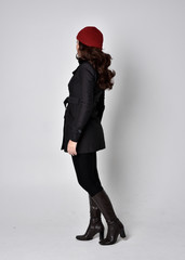 full length portrait of a pretty brunette girl wearing a black coat, leather boots and red beanie. Standing pose with back to the camera on a grey studio background.