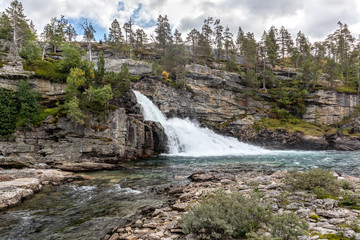 Fototapeta na wymiar Scenic waterfall on river in mountains of Norway with rocky wall and pine trees, wild nature northern landscape