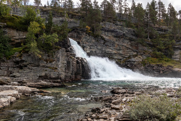 Scenic waterfall on river in mountains of Norway with rocky wall and pine trees, wild nature northern landscape