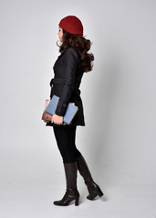 full length portrait of a pretty brunette girl wearing a black coat, leather boots and red beanie. Standing pose with back to the camera, holding books, on a grey studio background.