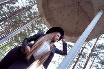 A young blonde woman in a black hat inside an architectural structure. A black leather jacket and a white shirt