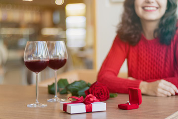 White giftbox, two wineglasses, a rose and a ring in the box laying on the table. Young woman wearing a red sweater smiling. Present with a red ribbon. Valentine's day concept.