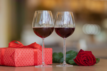 Red giftbox a rose and two glasses with wine on the table. Present with a red ribbon. Valentine's day and winter holidays gifts concept.