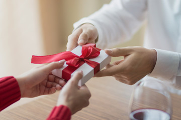 A small present is being passed by a couple from one to another. White little giftbox with a red ribbon in the hands. Valentine's day and winter holidays concept.