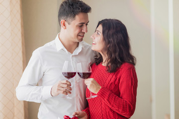 Young couple smiling to each other, drinking wine and holding white present with red ribbon in their hands at restaurant. Crossed wineglasses at a cafe, laugh and pass a gift. Valentine's day concept.
