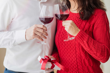 Young couple smiling to each other, drinking wine and holding white present with red ribbon in their hands at restaurant. Crossed wineglasses at a cafe, laugh and pass a gift. Valentine's day concept.