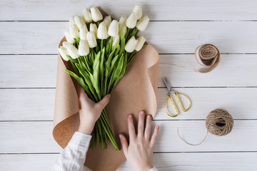 Woman florist wrapping beautiful bouquet of white tulips in pack craft paper on the wooden table. Flat lay