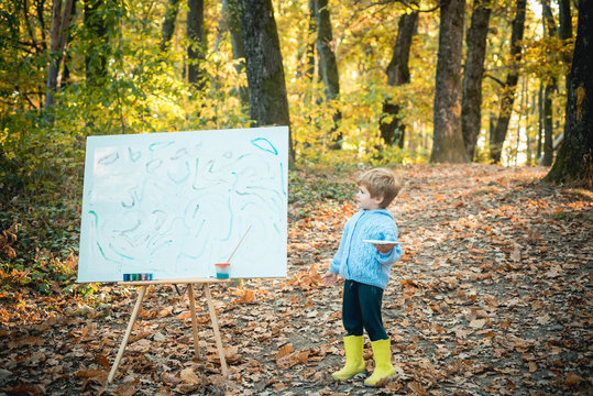Artwork of autumn baby boy in fall leaves park. Children creativity concept. Preschool boy with a brush paint on canvas at autumnal beautiful golden forest park. Art inspiration.
