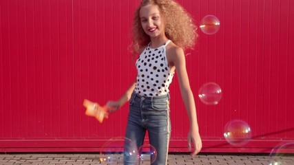 Curly blonde girl in a polka dot print t-shirt and blue jeans blows soap bubbles and winks against a red wall. Video