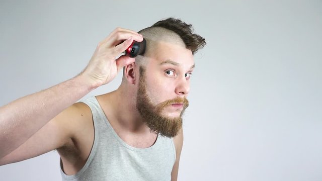 The guy shaves his head with clippers in front of a mirror.Bald and a beard.