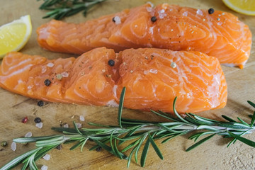 Two salmon or trout steaks lie on a wooden cutting board with fresh rosemary, chopped lemon, peppercorns and sea salt crystals. Horizontal orientation. Close-up.