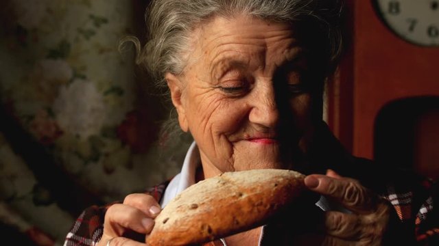 Elderly happy woman breaks off a piece of bread and eats on a dark background
