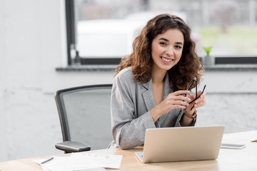 attractive and smiling account manager sitting at table and holding glasses