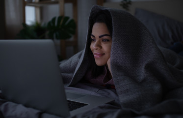 Beautiful young mixed race woman lying in bed at night, having a video call on a laptop computer.