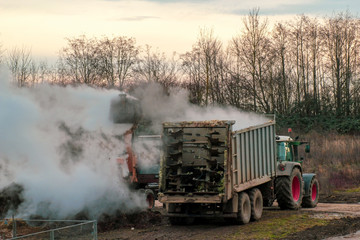 Excavator loads manure, hovering in the cold air, into a tractor with a trailer
