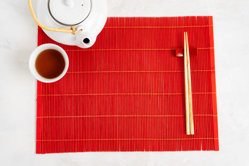 Asian tea food background. Teapot and Cup with chopsticks on the red bamboo mat over the gray stone background with copy space. Mockup for the menu.